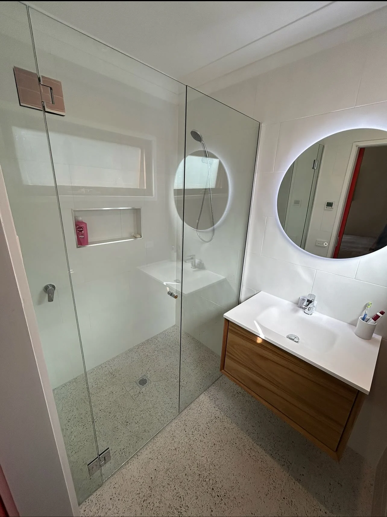 Completed bathroom with a floating timber vanity, round illuminated mirror and walk-in shower